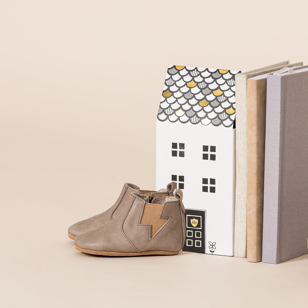 Side view of baby electric boots in colour taupe next to books and box shaped like house