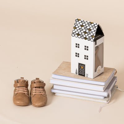 Baby Archie boots in colour Tan next to cardboard box shaped like a house