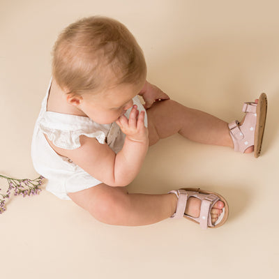 Baby sitting on ground wearing baby pink sandals with white daisy detail